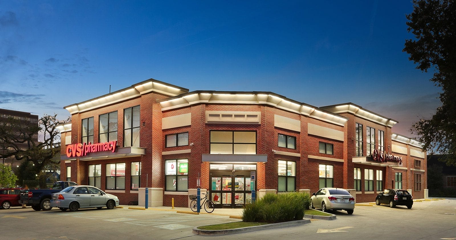 Exterior of a CVS Pharmacy store at twilight, featuring a brick building with illuminated signage and parked cars in the CVS Portfolio.