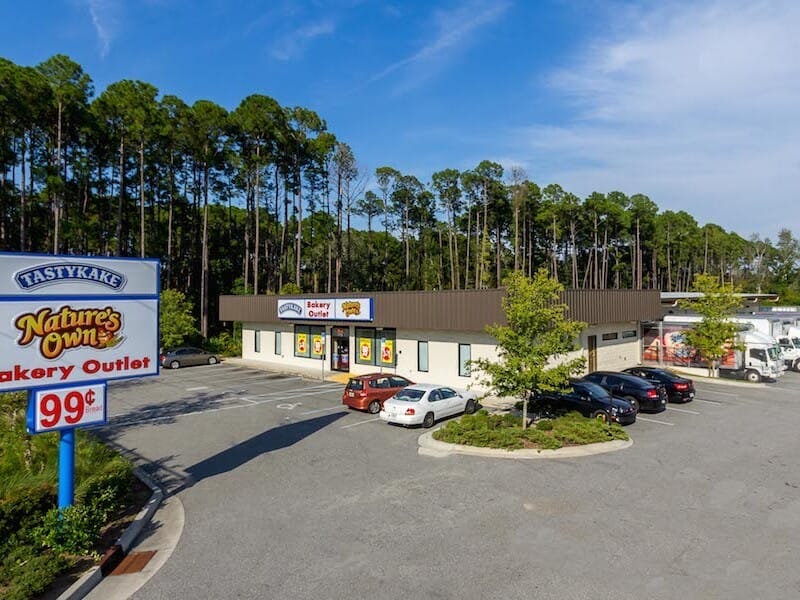 A small, single-story Flowers Foods bakery outlet surrounded by a parking lot with cars, located next to a wooded area under a clear sky.