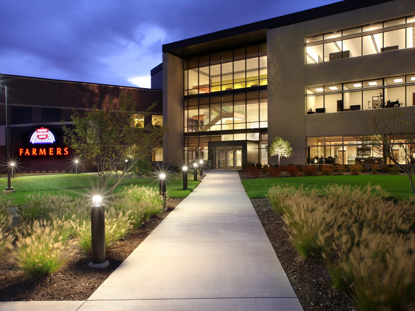Exterior view of a modern office building at dusk, featuring illuminated signage, well-lit walkways, and landscaped areas available for sale leasebacks.