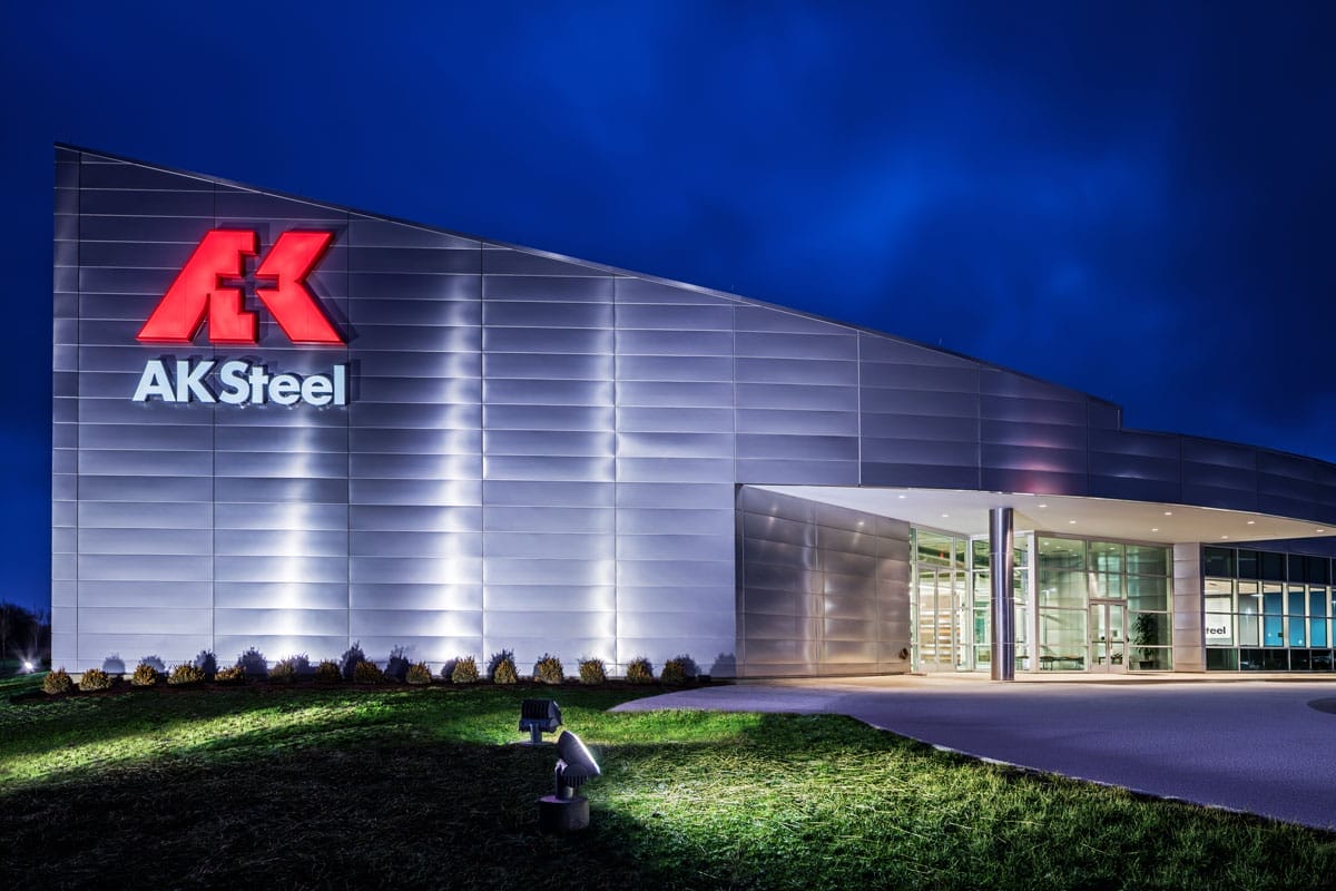 Exterior of the AK Steel building at night, featuring a brightly illuminated red ak logo on a modern silver facade, with a clear entrance and landscaped surroundings.