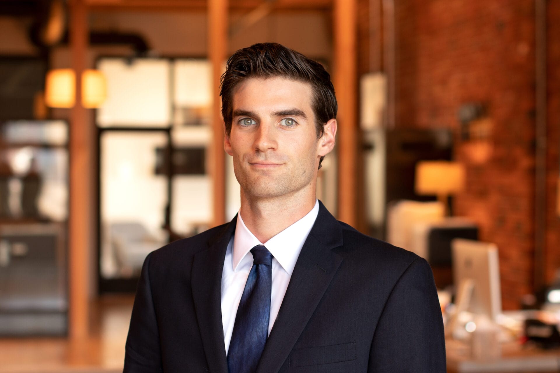 A professional man in a suit standing in an office with a brick wall background, recently hired by the Thomas Company.