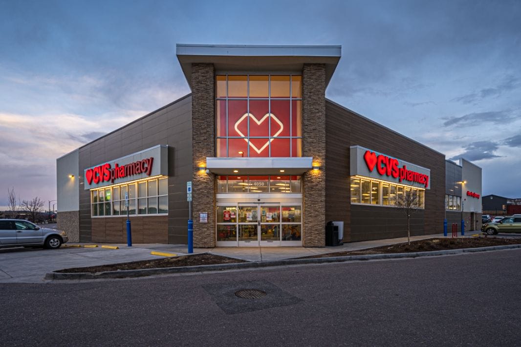 Exterior of a recently sold CVS pharmacy store in Castle Rock, Colorado at twilight with illuminated signage and parked cars under a cloudy sky.