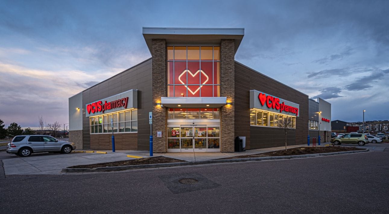 Exterior of a recently sold CVS pharmacy store in Castle Rock, Colorado at twilight with illuminated signage and parked cars under a cloudy sky.