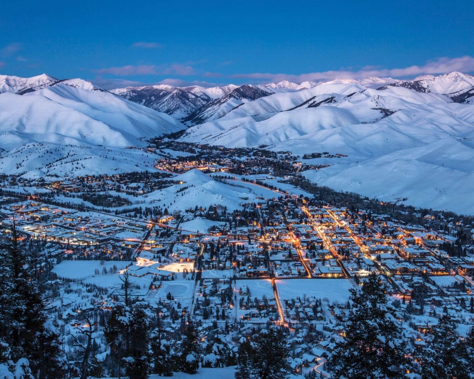 Aerial view of a snow-covered mountain town illuminated at dusk, with surrounding peaks under a twilight sky. For more information, contact us.