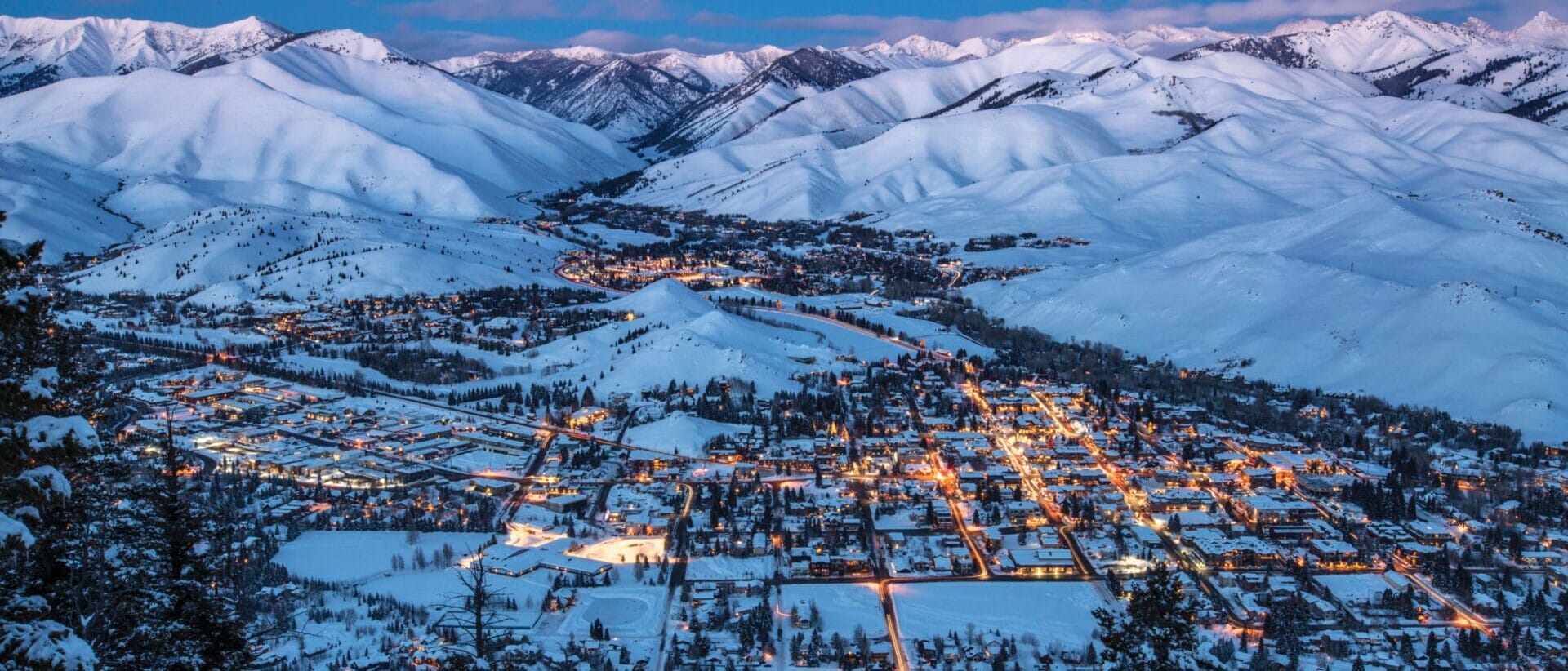 Aerial view of a snow-covered mountain town illuminated at dusk, with surrounding peaks under a twilight sky. For more information, contact us.