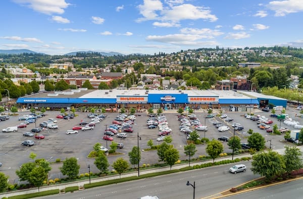 Aerial view of a bustling shopping center with McLendon Hardware, surrounded by parked cars and green hills in the background under a cloudy sky.