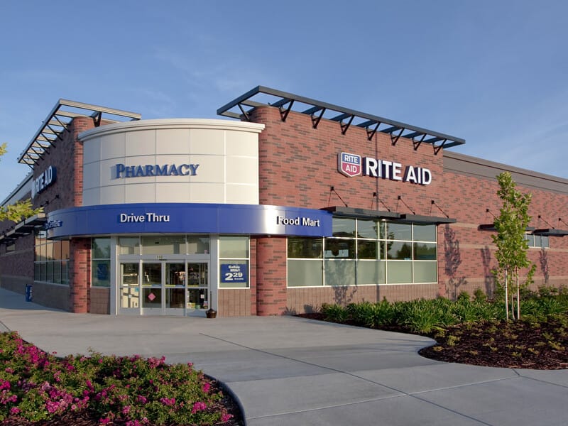 Exterior view of a Rite Aid drugstore with a drive-thru, set against a clear blue sky, surrounded by landscaped greenery.