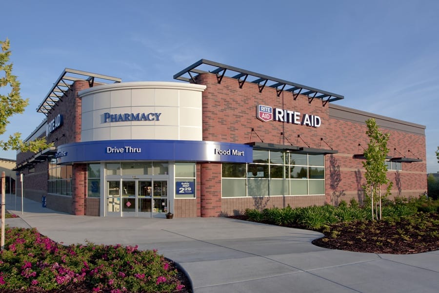 Exterior view of a Rite Aid drugstore with a drive-thru, set against a clear blue sky, surrounded by landscaped greenery.