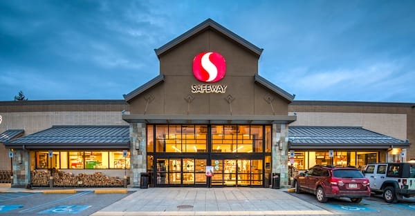 Exterior view of a Safeway grocery store in Lacey, WA at dusk, featuring illuminated signage, glass front doors, and parked cars outside.