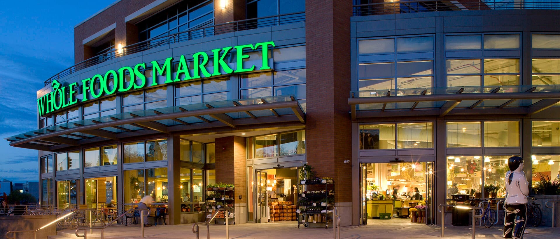 Exterior of a whole foods market at dusk with illuminated green signage, featuring a bicyclist at the entrance and visible interior lighting, exemplifying prime single tenant net leased investments nationwide.