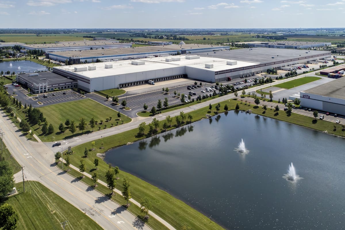 Aerial view of a large industrial distribution center with multiple warehouses, a parking lot, a small lake with fountains, and landscaped green areas.