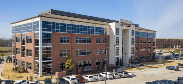 Aerial view of a modern multi-story office headquarters with a centenary logo, surrounded by a parking lot full of cars on a sunny day.