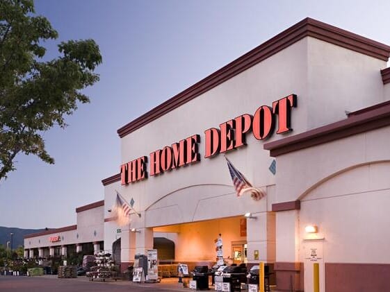 Exterior view of a Home Depot store at twilight, featuring the store's sign, a well-lit entrance, and a display of plants.