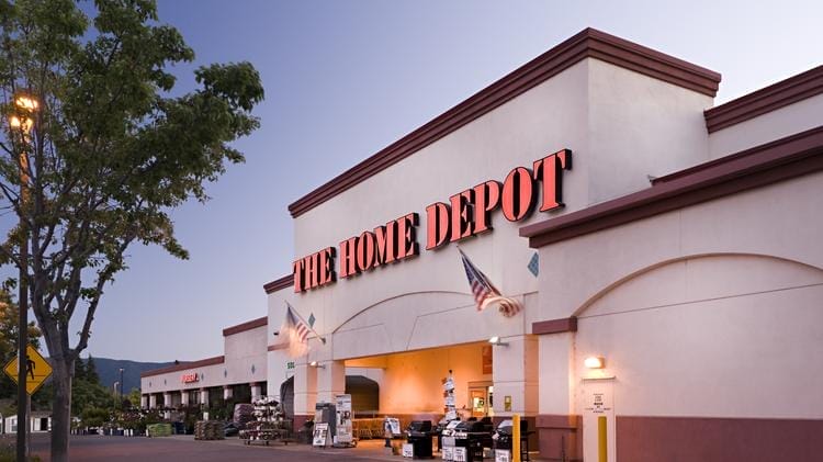 Exterior view of a Home Depot store at twilight, featuring the store's sign, a well-lit entrance, and a display of plants.