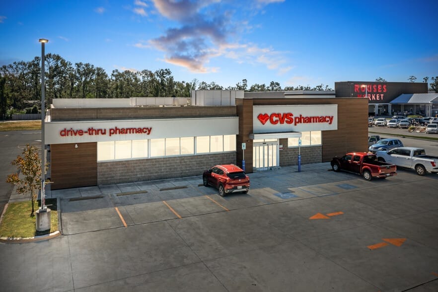 A recently sold CVS pharmacy building with a drive-thru pharmacy section, adjacent to a Rouses Market, under a cloudy sky in a parking lot with several cars in Moss Bluff, LA.