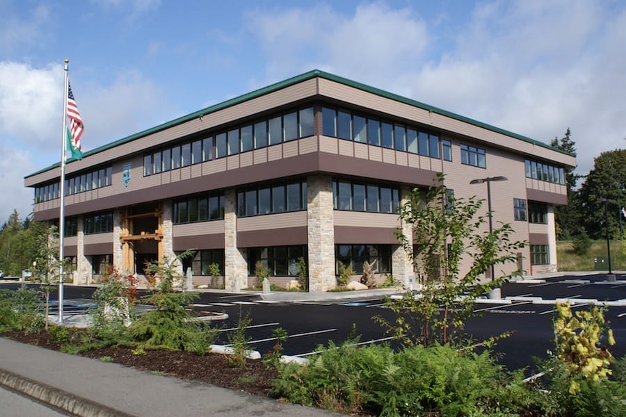 Modern two-story office building serving as a Washington State Parks facility, featuring a stone base and brown paneling, surrounded by a parking lot and green landscaping, under a clear sky.