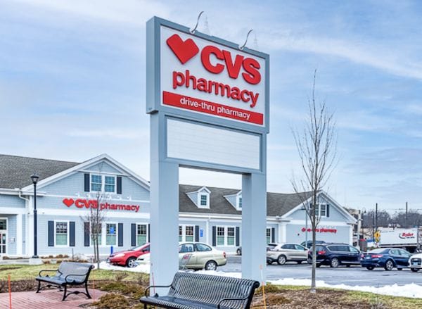 Signage for a CVS Pharmacy leasehold with a drive-thru, set against a backdrop of a clear blue sky and a building with several parked cars.