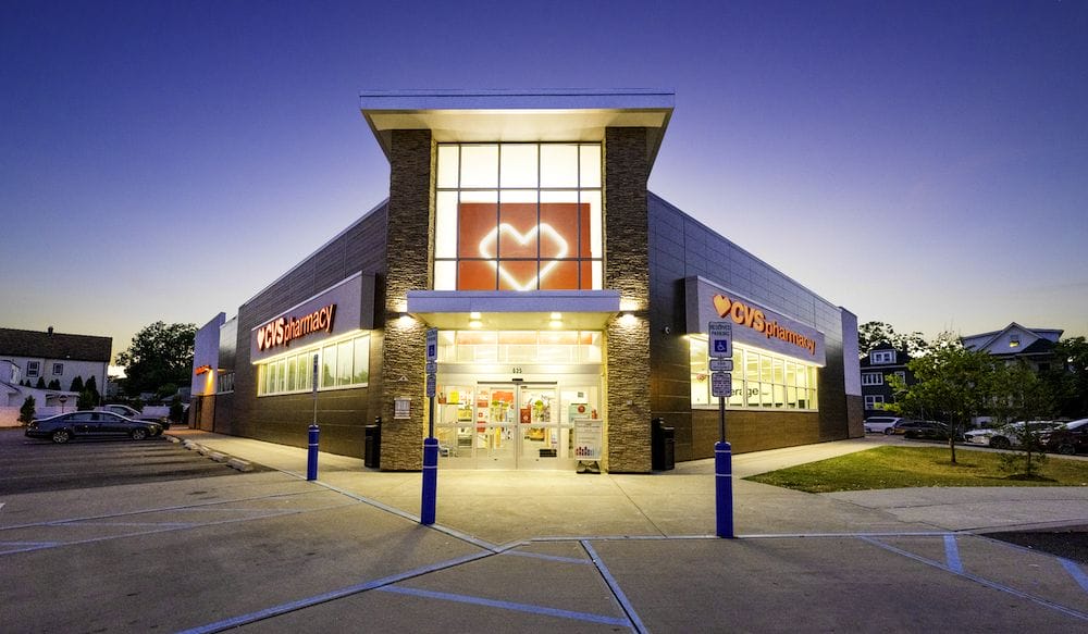 Exterior view of a CVS pharmacy store from a recent sale five-property CVS retail portfolio at twilight, featuring illuminated signage and clear glass doors, with a parking lot in the foreground.
