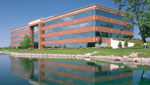 Modern multi-story HQ office building with reflective glass windows, situated beside a calm lake with a landscaped lawn and clear blue sky.
