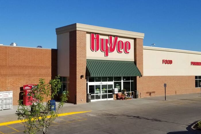 Exterior view of a Zero-Cash-Flow Hy-Vee grocery store on a sunny day, featuring prominent signage, a clear sky, and a small planted tree in the foreground.