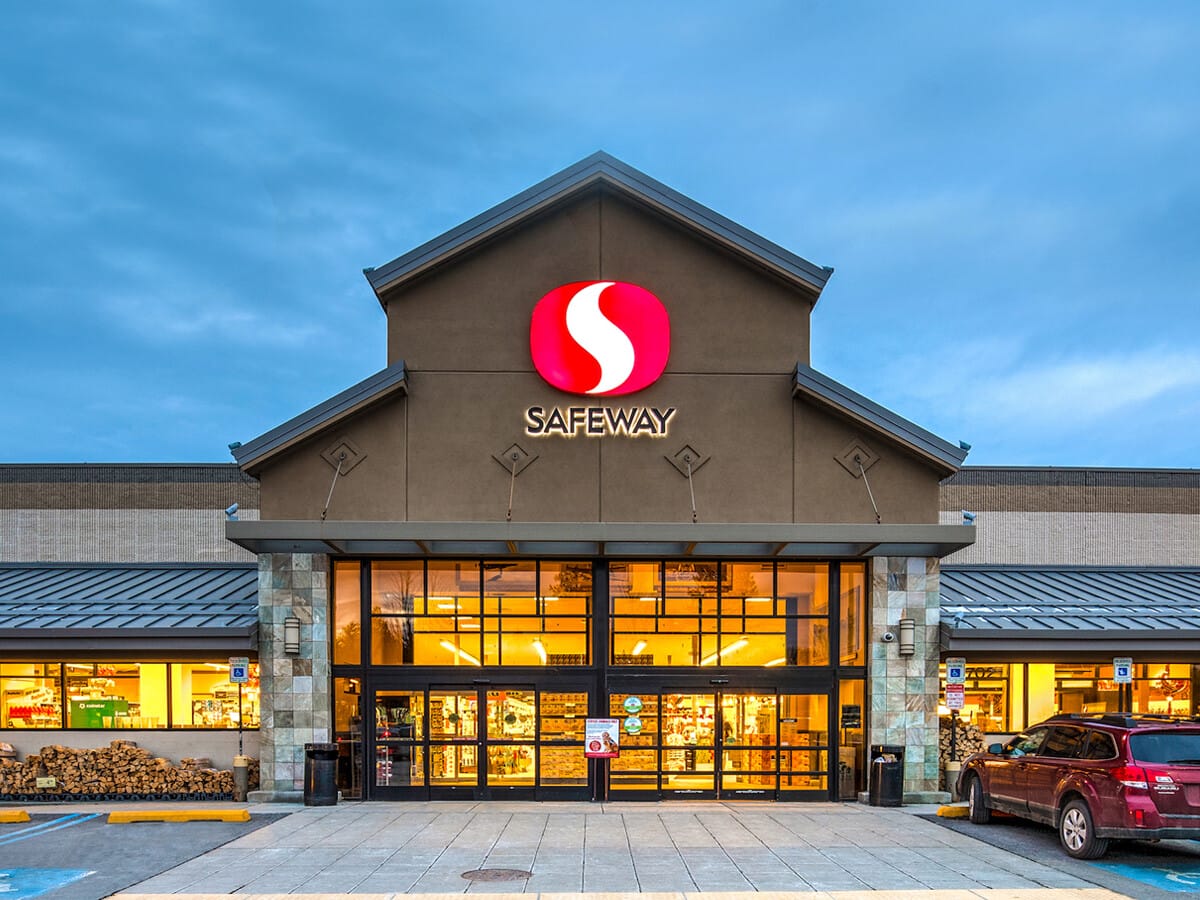 Exterior view of a Safeway grocery store at twilight with illuminated storefront and parked cars in front, representing single tenant net leased investments nationwide.