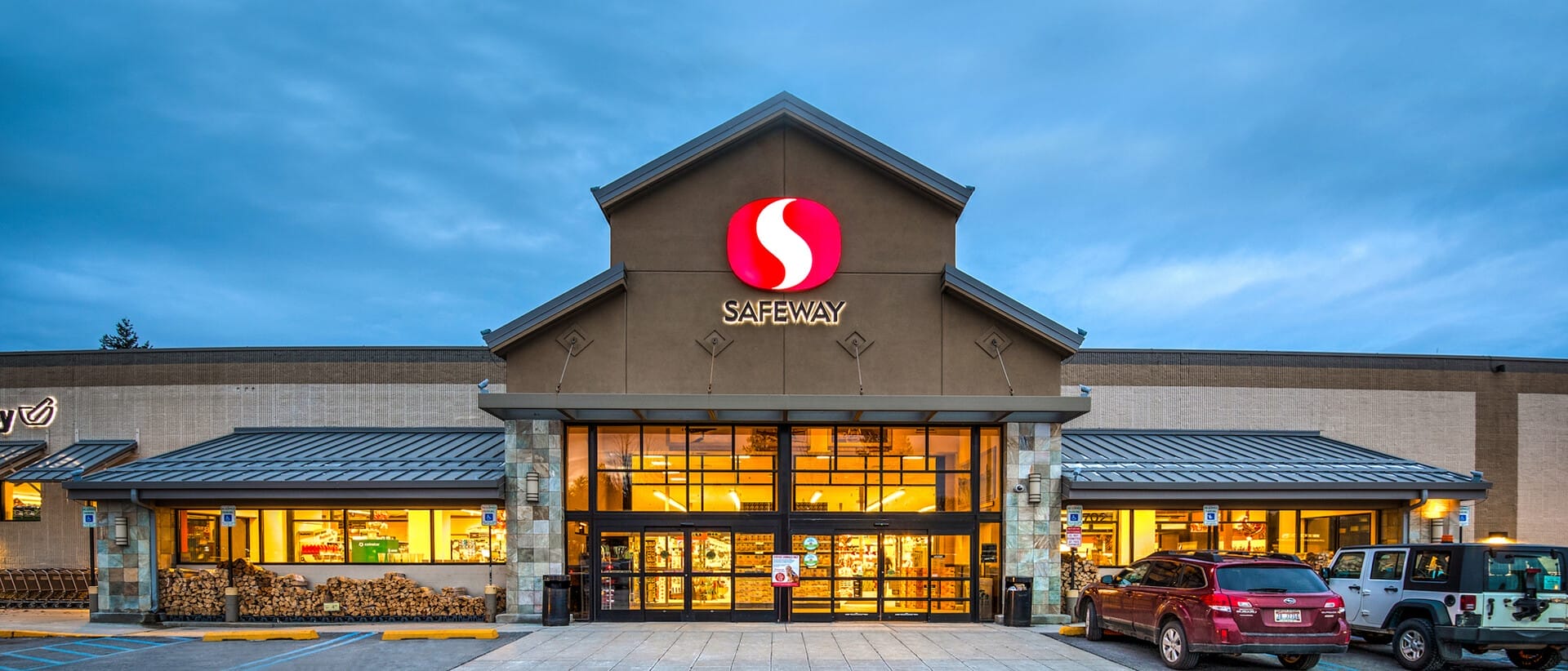 Exterior view of a Safeway grocery store at twilight with illuminated storefront and parked cars in front, representing single tenant net leased investments nationwide.