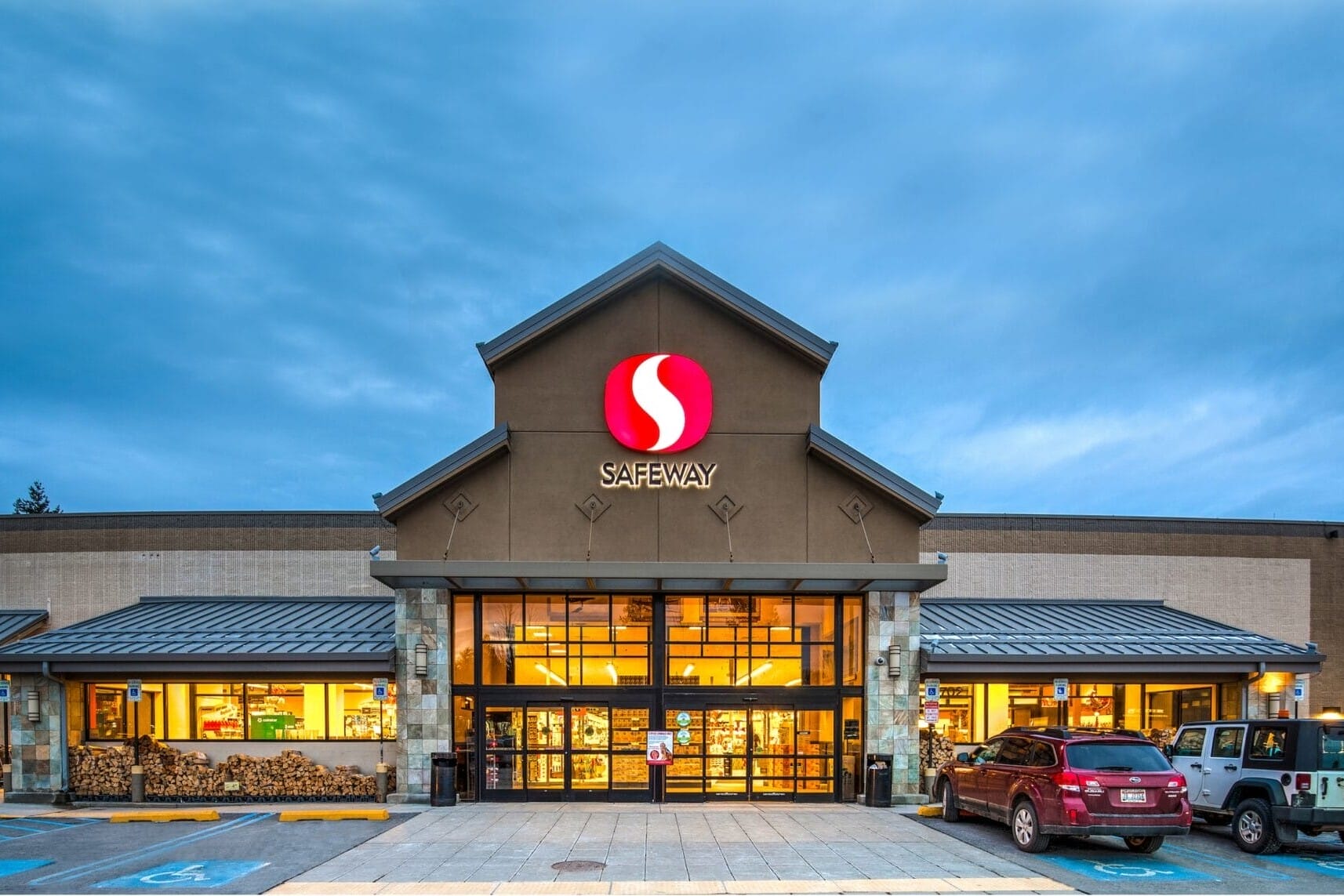 Exterior of a Safeway supermarket at dusk with illuminated signage and parked cars.