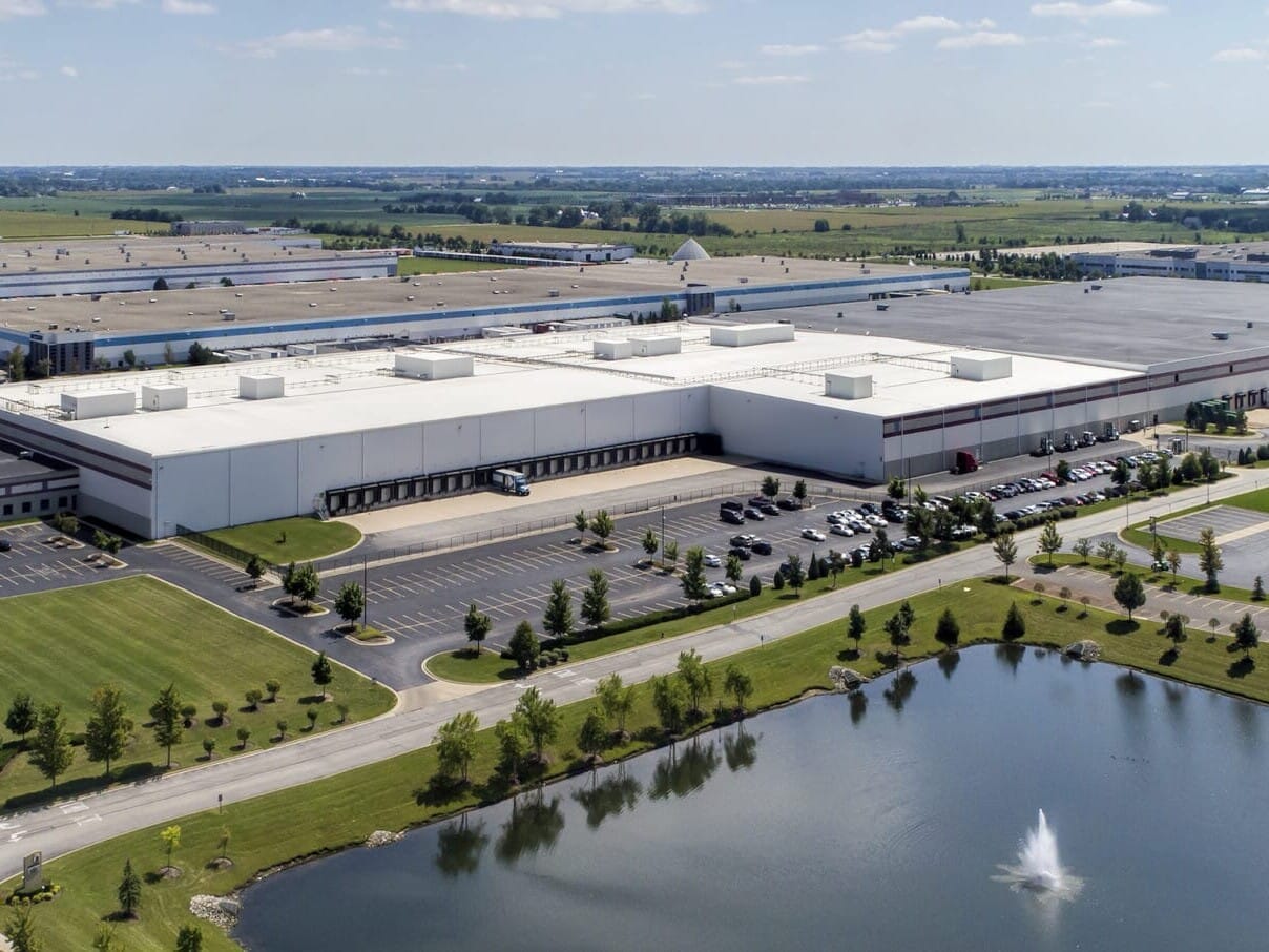 Aerial view of a large industrial park with multiple warehouses, parking spaces, and a pond with a fountain, specializing in single tenant net leased investments nationwide.