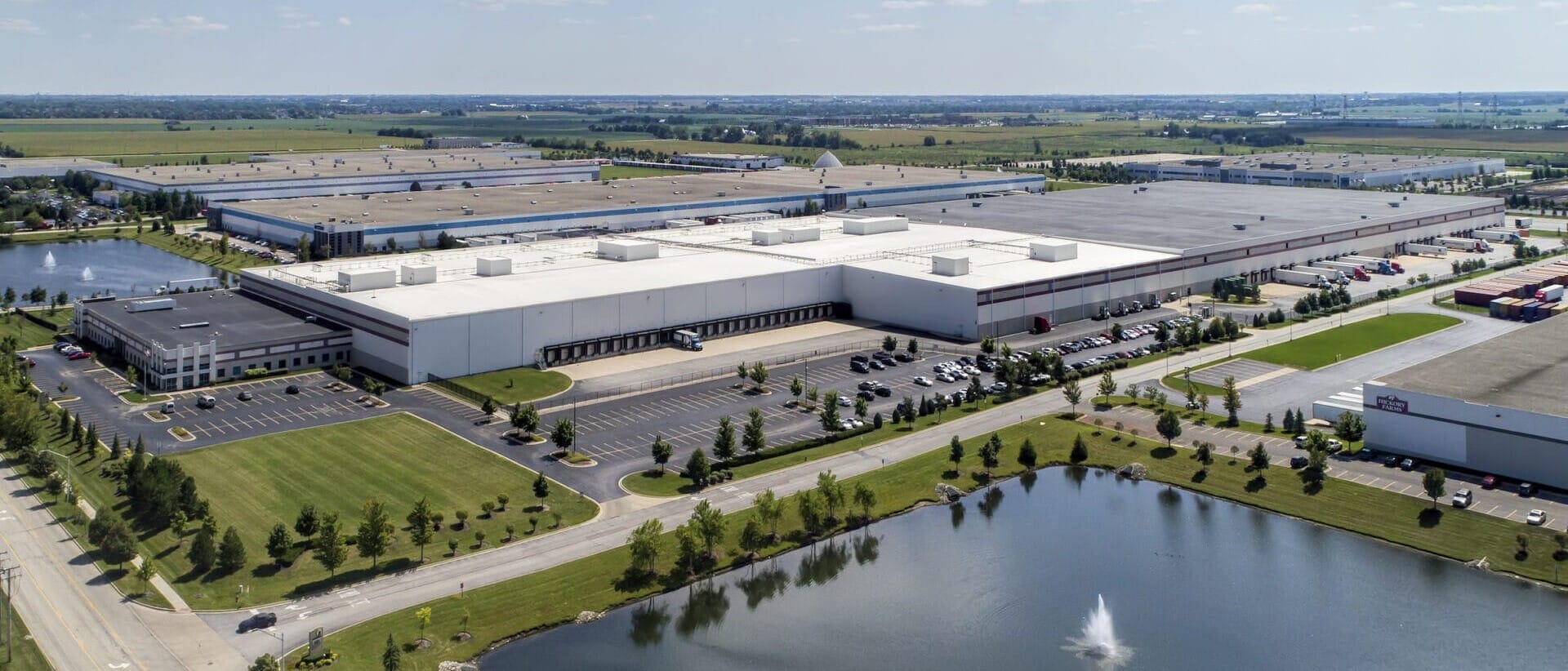 Aerial view of a large industrial park with multiple warehouses, parking spaces, and a pond with a fountain, specializing in single tenant net leased investments nationwide.