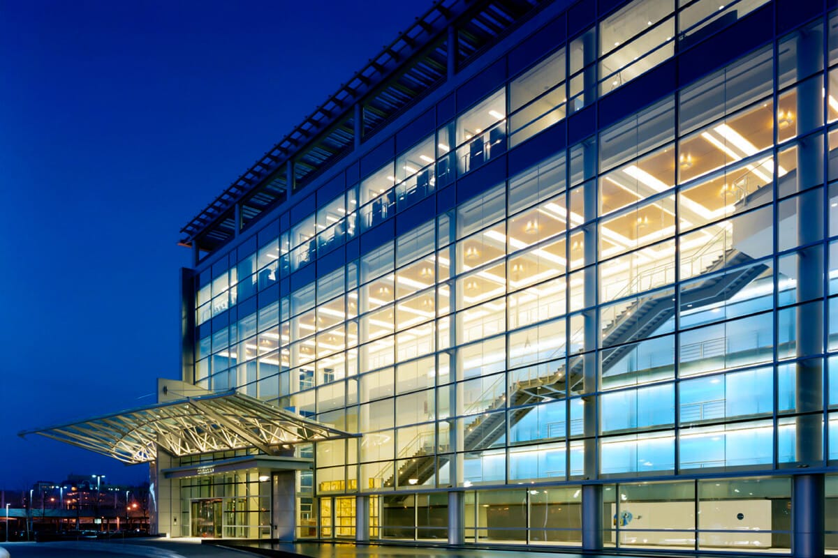 Modern glass building illuminated at twilight, representing a prime example of real estate investments, featuring a large transparent facade with visible interior staircases and exterior metal framework.