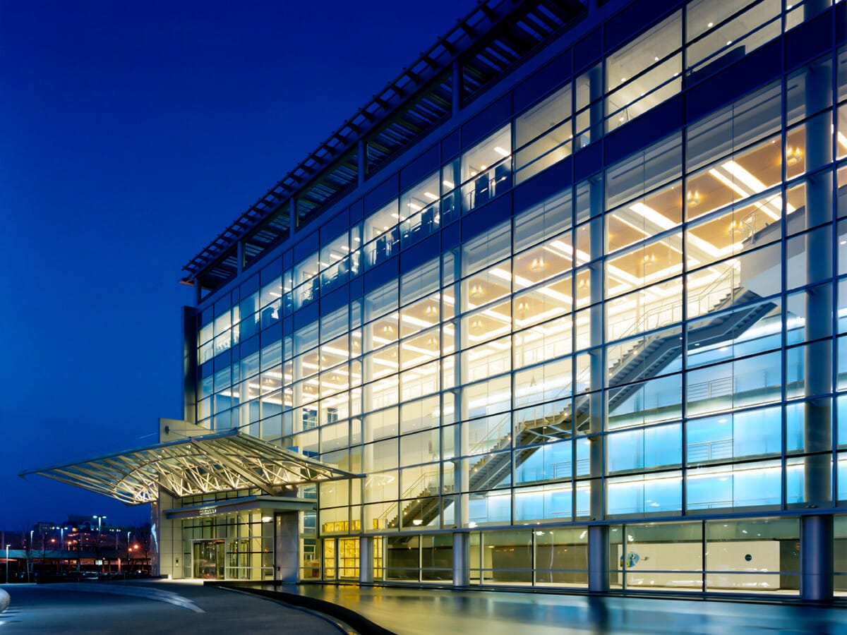 Modern glass building illuminated at twilight, representing a prime example of real estate investments, featuring a large transparent facade with visible interior staircases and exterior metal framework.