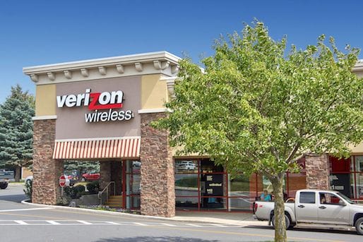 Exterior view of a recently sold Verizon Wireless store in a suburban shopping plaza with parked cars and trees under a clear sky.