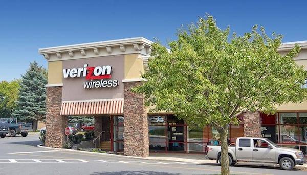 Exterior view of a recently sold Verizon Wireless store in a suburban shopping plaza with parked cars and trees under a clear sky.