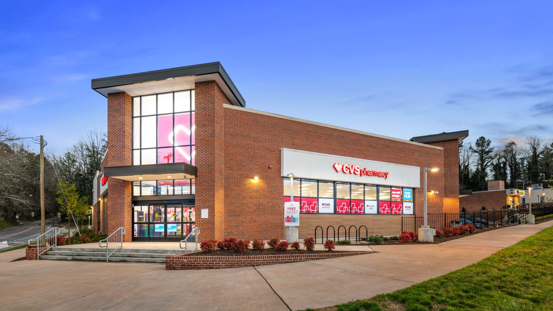 A brick CVS Pharmacy building with a large window at the entrance, illuminated at dusk. The sign reads 