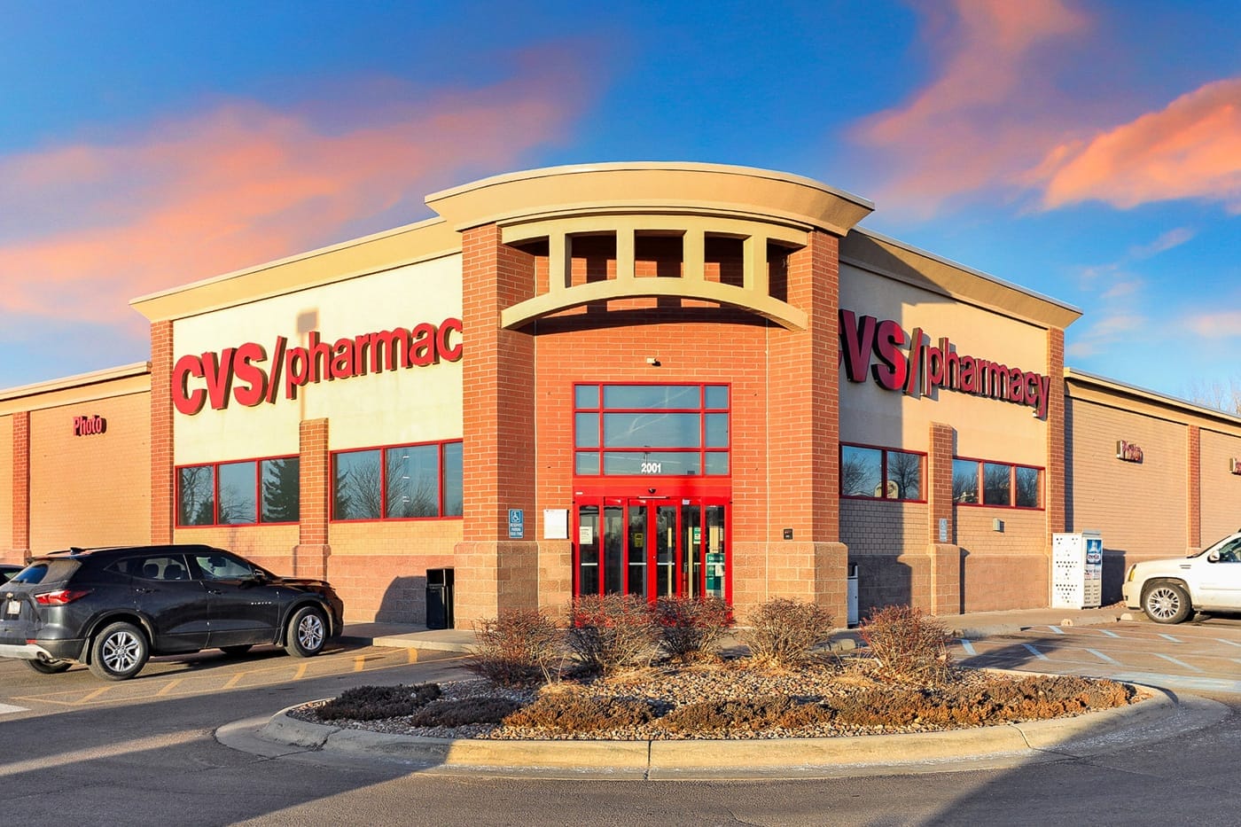 A CVS Pharmacy storefront with red signage on a brick building, cars parked outside, and a blue sky with clouds in the background—part of a recently sold 2-property CVS portfolio.