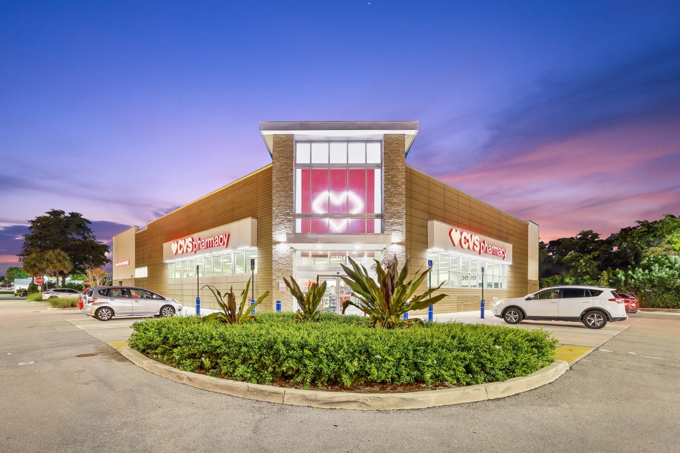 A CVS Pharmacy storefront in Miami at dusk, with illuminated signage, parked cars, and a landscaped area in front—this location was recently sold.