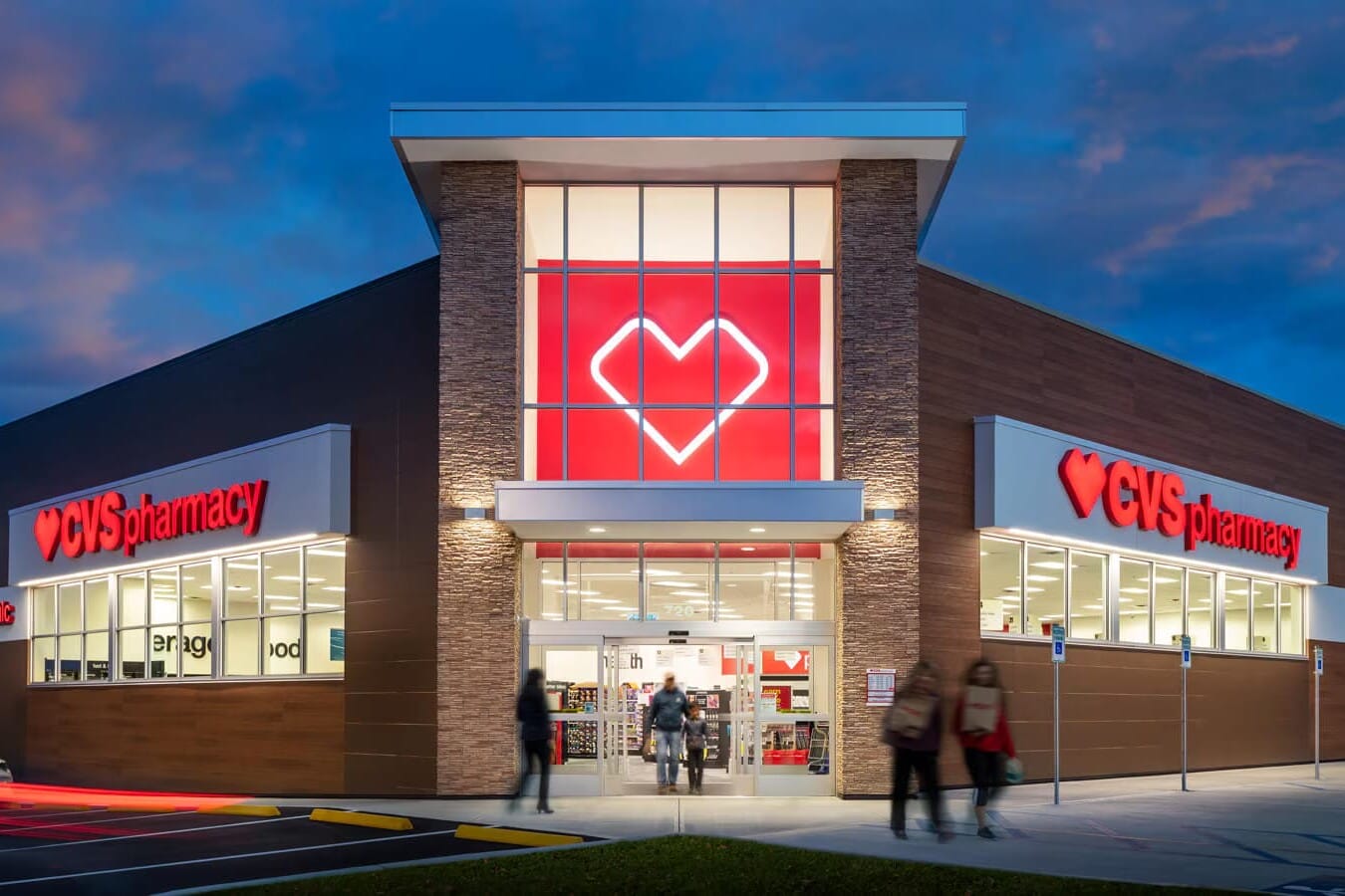 Exterior view of a recently sold CVS Pharmacy store at dusk with illuminated signage, large glass entrance, and several people entering and exiting.